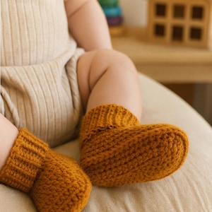 Peut inclure: Bottes de bébé au crochet jaune moutarde. Les chaussons sont faits d'un fil texturé et sont présentés sur les pieds d'un bébé. Le bébé porte un body beige. Une maison en bois et un jouet d'empilage arc-en-ciel sont en arrière-plan.