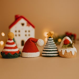 Puede incluir: Cuatro adornos navideños de ganchillo, incluyendo un gorro de Papá Noel, un árbol de Navidad y un pastel. Los adornos son rojos, blancos, verdes y amarillos. Una casa de cerámica blanca está en el fondo.