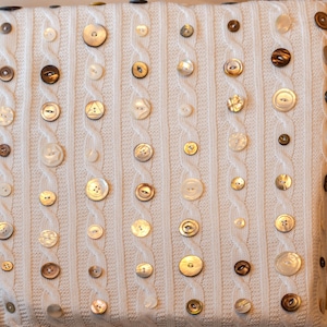 May include: A square, off-white pillow with a cable knit design, adorned with various sizes and colors of buttons. The buttons are in shades of white, cream, and brown, adding texture and visual interest to the decorative pillow.