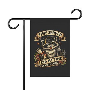 May include: Black garden flag with a raccoon illustration wearing a graduation cap and holding a scroll. Text reads "TIME SERVED", "I DID MY TIME CLASS OF 2026". The raccoon is winking and giving a thumbs up.