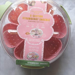 Peut inclure: Un récipient à collation rose en forme de fraise avec six sections amovibles. Le couvercle transparent et la poignée verte sont visibles. L'emballage indique "6-SECTION STRAWBERRY SNACKLE" et est conçu pour stocker fruits, légumes, bonbons, trempettes et tartinades.