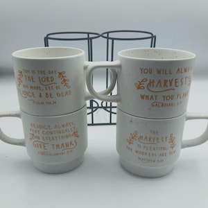 Puede incluir: Cuatro tazas de cerámica blanca moteada apiladas en pares. Cada taza tiene un asa curva y presenta texto inspirador en naranja. Un estante de metal negro está en el fondo. Las tazas son un producto de temática religiosa.