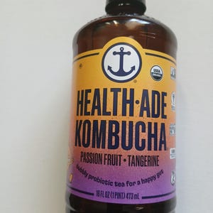 Może przedstawiać: Brązowa szklana butelka Health-Ade Kombucha z czarną zakrętką. Etykieta zawiera logo kotwicy i napis "Passion Fruit - Tangerine". Butelka zawiera 473 ml musującej herbaty probiotycznej.