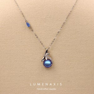 May include: A silver necklace with a blue iridescent pearl pendant. The pendant is accented with a dragonfly design and small clear stones. A small blue teardrop-shaped stone is set on the chain. The brand name "LUMENAXIS" is printed at the bottom.