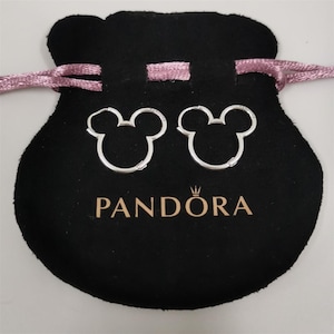 Puede incluir: Un par de pendientes de aro plateados con forma de cabeza de Mickey Mouse, presentados en una bolsa de terciopelo negro con el logotipo "PANDORA" y un cord&oacute;n rosa.