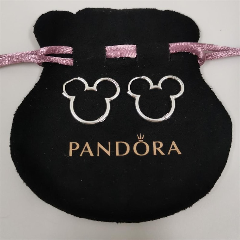 Puede incluir: Un par de pendientes de aro plateados con forma de cabeza de Mickey Mouse, presentados en una bolsa de terciopelo negro con el logotipo "PANDORA" y un cord&oacute;n rosa.