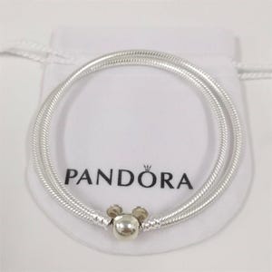 Puede incluir: Una pulsera Pandora plateada con un charm de Mickey Mouse. La pulsera tiene un dise&ntilde;o de doble hebra con un patr&oacute;n de cadena de serpiente texturizada. El charm es una esfera plateada con dos peque&ntilde;as orejas redondas plateadas. La pulsera est&aacute; sobre una bolsa blanca con el logo de Pandora.