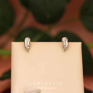 Puede incluir: Un par de pendientes de plata con forma de pluma, cada uno con una pequeña gema transparente. Los pendientes se exhiben sobre una superficie de tono neutro contra un fondo borroso verde y marrón. La marca "LUMENAXIS" y el lema "Handcrafted Jeweller" son visibles.