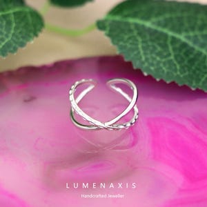 May include: A silver-colored adjustable ring with an X-shaped design. The ring has a textured pattern on the band and is displayed on a pink surface with green leaves in the background. The brand name "LUMENAXIS" is visible.