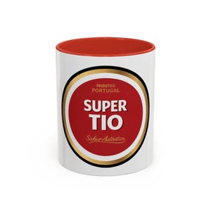 Puede incluir: Una taza de cerámica blanca con interior y borde rojos. El frente presenta un logotipo circular con fondo rojo, bordes dorados y negros, y el texto blanco SUPER TIO. Arriba del texto dice PRODUTO DE PORTUGAL y debajo Sabor Autêntico.