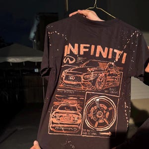 May include: A black short-sleeve t-shirt with a bleached splatter effect and a graphic print in orange. The design features an Infiniti car, the text "INFINITI", "STAY INSPIRED", "BUILT NOT BOUGHT", and "TO INFINITY AND BEYOND" with a car wheel.