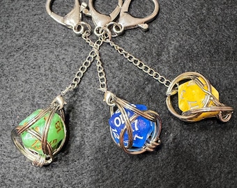 May include: Three dice keychains with silver-colored metal hardware. Each die is a different color: green, blue, and yellow. The dice are held in place by a wire cage and attached to a chain and a clasp. The keychains are on a dark gray surface.