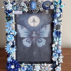 May include: A rectangular picture frame with a butterfly print. The frame is embellished with various blue and silver decorative elements, including flowers, beads, and cameos. The frame is made of metal and has a glass front.