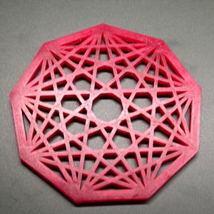 May include: A red, geometric coaster with an intricate, openwork design. The coaster is octagonal with a central circular opening and a pattern of intersecting lines and shapes. The coaster is made of a solid material and sits on a gray surface.