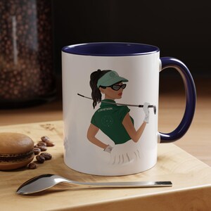 May include: White ceramic mug with a navy blue interior and handle. The mug features an illustration of a woman in a golf outfit, wearing sunglasses and a visor, holding a golf club. The mug is on a wooden surface.