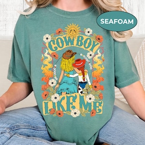 Puede incluir: Una camiseta verde espuma de mar con una ilustraci&oacute;n gr&aacute;fica de dos figuras con sombreros de vaquero, cabello rubio y pelirrojo, rodeadas de flores, llamas y un sol. El texto dice COWBOY LIKE ME, con un lazo formando un c&iacute;rculo alrededor.