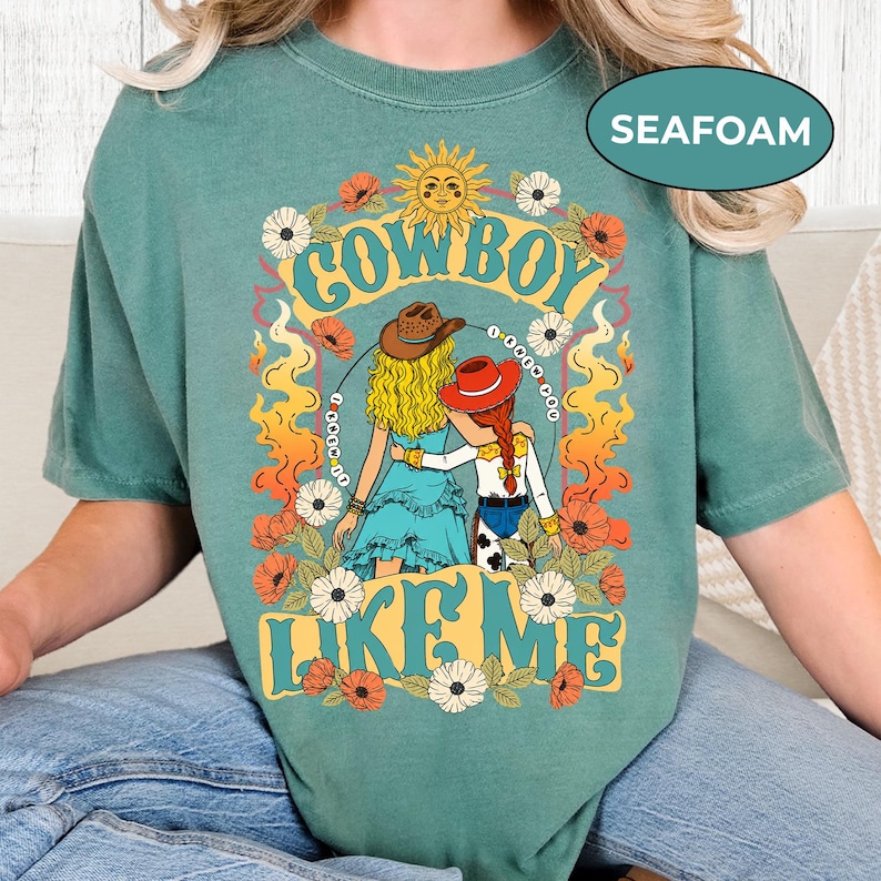 Puede incluir: Una camiseta verde espuma de mar con una ilustraci&oacute;n gr&aacute;fica de dos figuras con sombreros de vaquero, cabello rubio y pelirrojo, rodeadas de flores, llamas y un sol. El texto dice COWBOY LIKE ME, con un lazo formando un c&iacute;rculo alrededor.