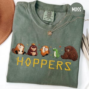 May include: Moss green t-shirt with embroidered cartoon animals and the word "HOPPERS" in yellow. The shirt features a beaver, a bear, a frog, and other woodland creatures. The shirt is a Comfort Colors brand.