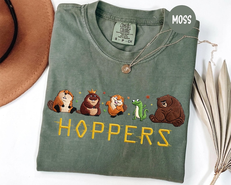 May include: Moss green t-shirt with embroidered cartoon animals and the word "HOPPERS" in yellow. The shirt features a beaver, a bear, a frog, and other woodland creatures. The shirt is a Comfort Colors brand.