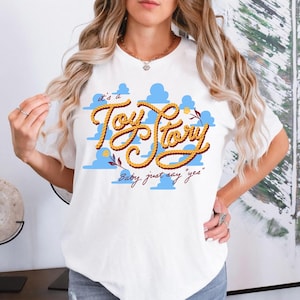May include: A white short-sleeved t-shirt featuring a graphic print of light blue clouds and the text "it's a Toy Story" written in a yellow rope-style font, with the additional text "Baby, just say "yes"" printed in a dark red cursive script.