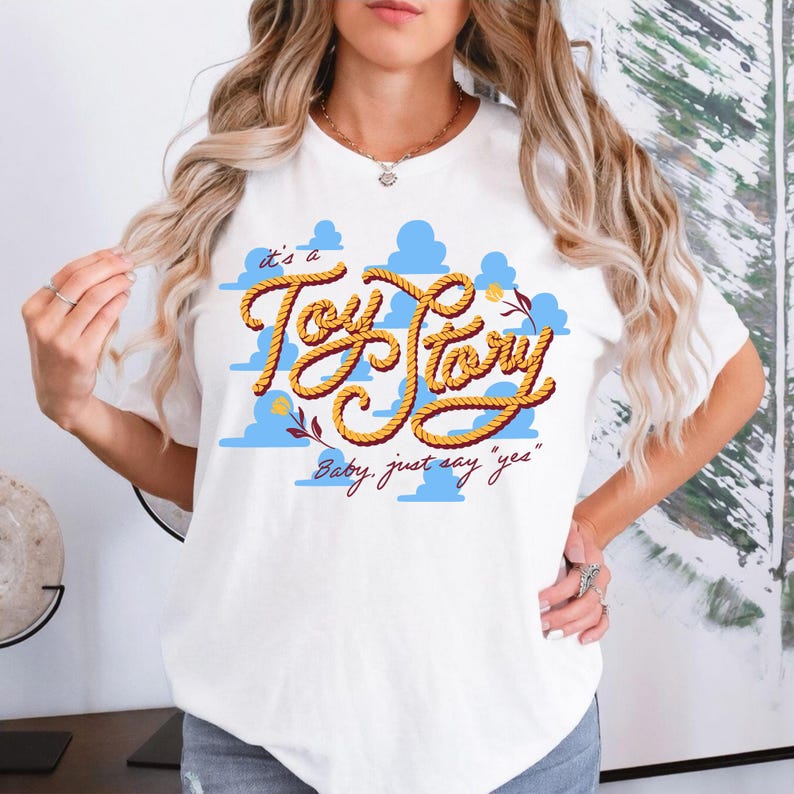 May include: A white short-sleeved t-shirt featuring a graphic print of light blue clouds and the text "it's a Toy Story" written in a yellow rope-style font, with the additional text "Baby, just say "yes"" printed in a dark red cursive script.