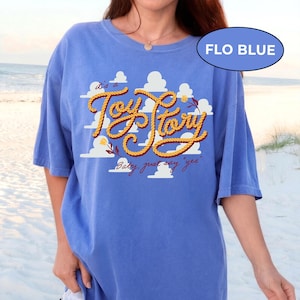 May include: A periwinkle blue t-shirt featuring a graphic print of white clouds and the text "it's a Toy Story" written in a yellow rope-style font, with the additional text "Baby, just say 'yes'" below and a small flower accent.