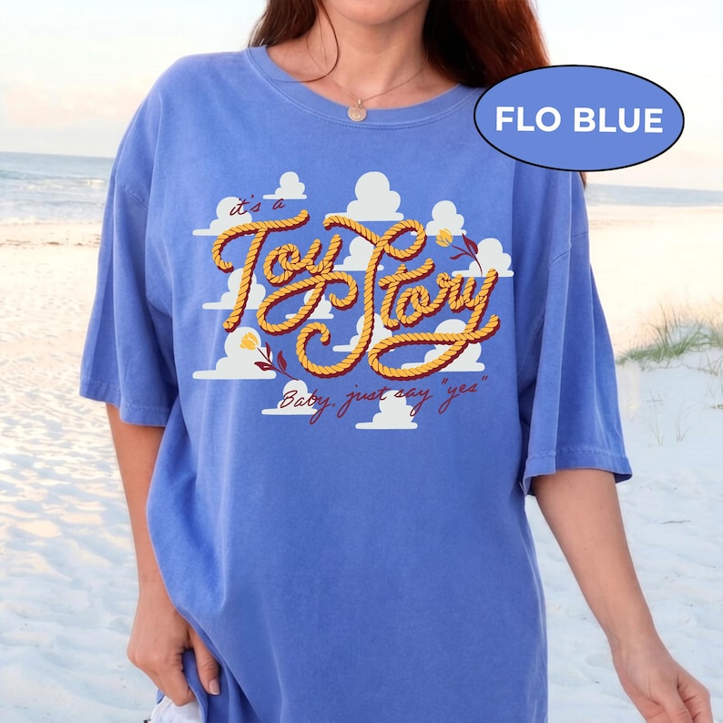 May include: A periwinkle blue t-shirt featuring a graphic print of white clouds and the text "it's a Toy Story" written in a yellow rope-style font, with the additional text "Baby, just say 'yes'" below and a small flower accent.