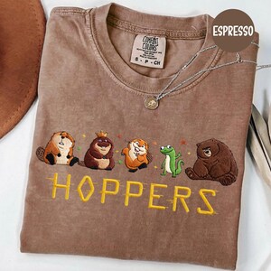 May include: Espresso-colored t-shirt with embroidered cartoon animals and the word "HOPPERS" in yellow. The shirt features a beaver, a bear with a crown, an otter, a frog, and a bear. A silver necklace with a pendant is on the shirt.