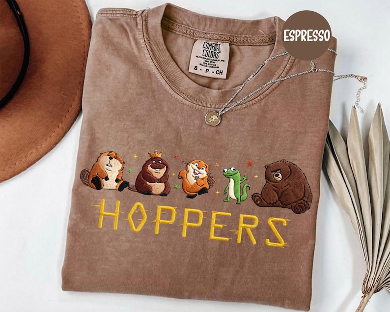 May include: Espresso-colored t-shirt with embroidered cartoon animals and the word "HOPPERS" in yellow. The shirt features a beaver, a bear with a crown, an otter, a frog, and a bear. A silver necklace with a pendant is on the shirt.