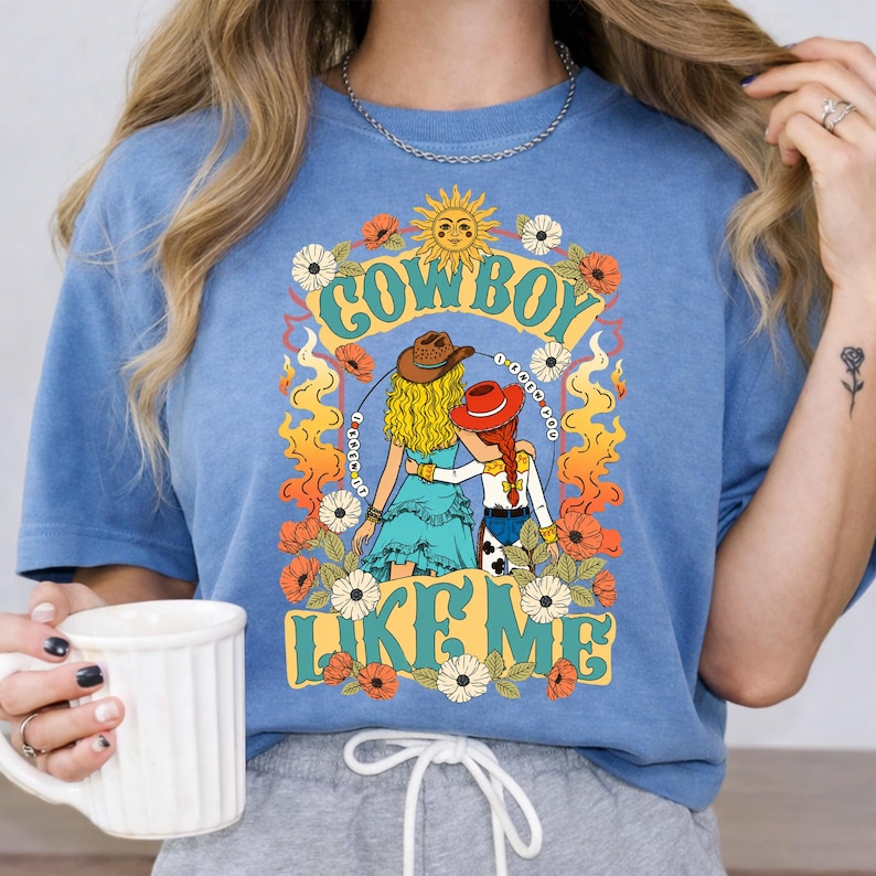 Puede incluir: Una camiseta azul de manga corta con un estampado gr&aacute;fico de dos figuras con sombreros de vaquero, una con cabello rubio y otra con cabello rojo, rodeadas de llamas naranjas y flores, con el texto "COWBOY LIKE ME" en una fuente retro en negrita.