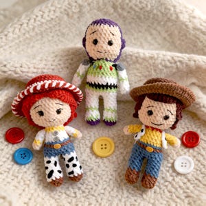 May include: Three handmade crochet dolls of characters from the Toy Story movie. The dolls include Jessie, Buzz Lightyear, and Woody. The dolls are surrounded by colorful buttons. The dolls are made of yarn and have embroidered facial features.