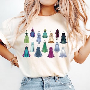 May include: Cream-colored t-shirt featuring a collection of illustrated dresses in various colors and styles. The dresses are arranged in rows, showcasing a range of designs, from ball gowns to more casual attire. The overall aesthetic is whimsical and artistic.