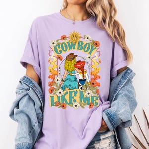 Puede incluir: Una camiseta de color morado claro con un estampado gr&aacute;fico de dos figuras con sombreros de vaquero y el texto COWBOY LIKE ME. El dise&ntilde;o incluye un sol, flores y llamas en tonos amarillos, naranjas y azules. Se lleva con una chaqueta vaquera.