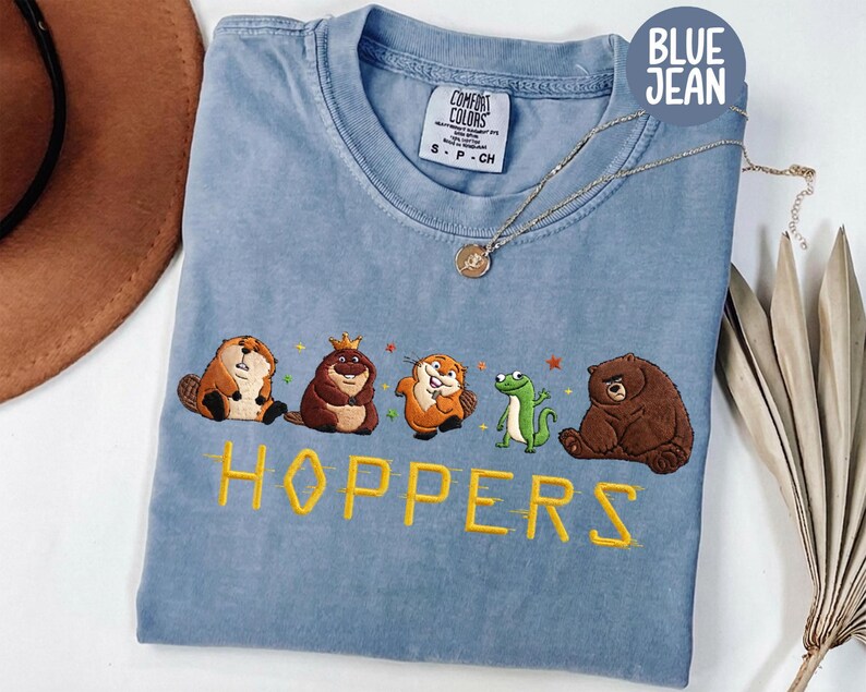 May include: Blue jean colored t-shirt with embroidered cartoon animals and the word "HOPPERS" in yellow. The shirt features a beaver, a bear, a frog, and other animals. A gold necklace is draped on the shirt.