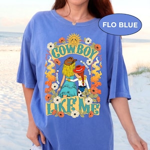 Puede incluir: Una camiseta azul b&iacute;garo con un estampado gr&aacute;fico que incluye el texto "COWBOY LIKE ME". El dise&ntilde;o muestra dos figuras con cabello rubio y rojo, usando sombreros, rodeadas de llamas naranjas, un sol amarillo y flores blancas y naranjas.