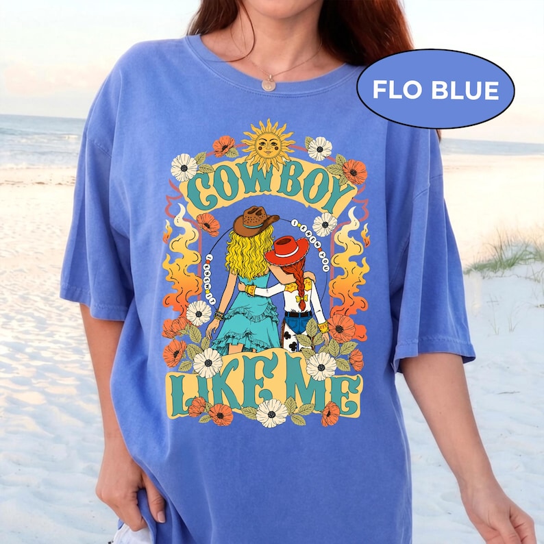 Puede incluir: Una camiseta azul b&iacute;garo con un estampado gr&aacute;fico que incluye el texto "COWBOY LIKE ME". El dise&ntilde;o muestra dos figuras con cabello rubio y rojo, usando sombreros, rodeadas de llamas naranjas, un sol amarillo y flores blancas y naranjas.