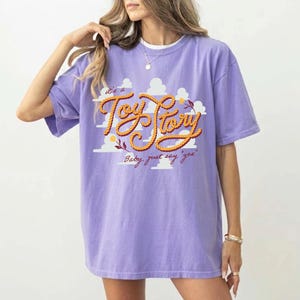 May include: A light purple t-shirt featuring a graphic print of white clouds and the text "It's a Toy Story" in a yellow rope-style font, with the additional script text "Baby, just say 'yes'" printed in a dark red cursive font below the main graphic.