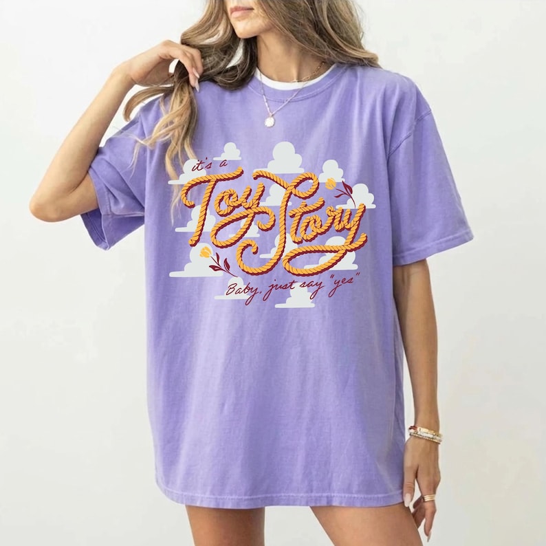 May include: A light purple t-shirt featuring a graphic print of white clouds and the text "It's a Toy Story" in a yellow rope-style font, with the additional script text "Baby, just say 'yes'" printed in a dark red cursive font below the main graphic.