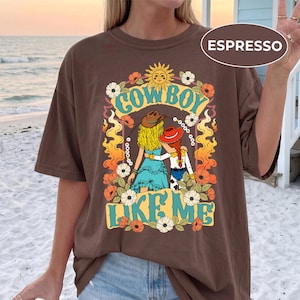 Puede incluir: Una camiseta de color marr&oacute;n oscuro con un estampado gr&aacute;fico de dos figuras con cabello rubio y rojo que llevan sombreros de vaquero. El dise&ntilde;o incluye el texto "COWBOY LIKE ME" en amarillo, rodeado de llamas naranjas, flores blancas y un sol.