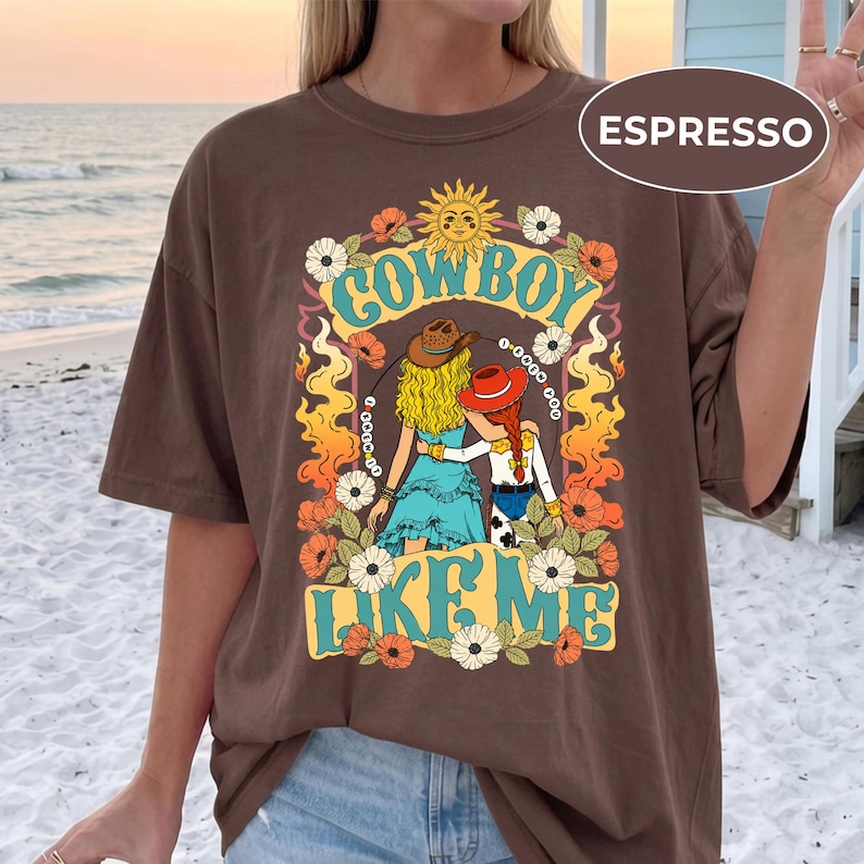 Puede incluir: Una camiseta de color marr&oacute;n oscuro con un estampado gr&aacute;fico de dos figuras con cabello rubio y rojo que llevan sombreros de vaquero. El dise&ntilde;o incluye el texto "COWBOY LIKE ME" en amarillo, rodeado de llamas naranjas, flores blancas y un sol.