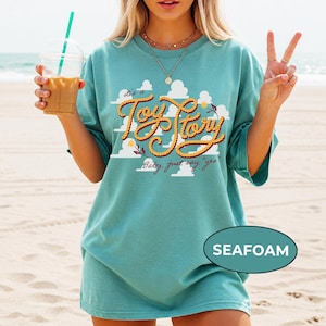 May include: A seafoam green short-sleeve t-shirt featuring a graphic of white clouds and the text "it's a Toy Story" written in a yellow rope-style font, with the additional text "Baby, just say "yes"" printed below in a cursive script.