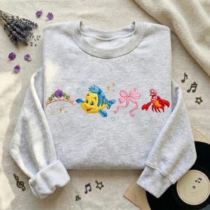 May include: A light gray sweatshirt with embroidered images of a purple crown, a yellow and blue fish, a pink bow, and a red crab. The sweatshirt has long sleeves and a crew neck.