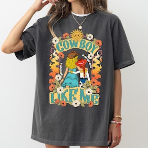 Puede incluir: Una camiseta de manga corta gris carb&oacute;n con un estampado gr&aacute;fico de dos figuras con sombreros de vaquero, una con cabello rubio y otra con trenzas rojas, rodeadas de flores, llamas, un sol y el texto "COWBOY LIKE ME" en una fuente azul claro.