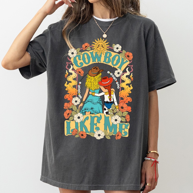 Puede incluir: Una camiseta de manga corta gris carb&oacute;n con un estampado gr&aacute;fico de dos figuras con sombreros de vaquero, una con cabello rubio y otra con trenzas rojas, rodeadas de flores, llamas, un sol y el texto "COWBOY LIKE ME" en una fuente azul claro.