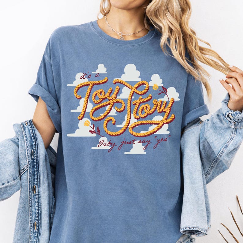 May include: A blue t-shirt featuring a graphic of white clouds and the text "it's a Toy Story" written in a yellow rope-style font, with the additional text "Baby, just say "yes"" in red cursive script and small yellow flower illustrations.