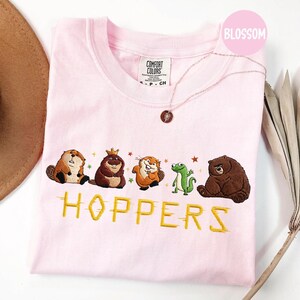 May include: A light pink Comfort Colors crew neck t-shirt featuring embroidered cartoon animals and the word "HOPPERS" in yellow. The animals include a beaver, a bear with a crown, a squirrel, a frog, and a brown bear. A gold necklace is also visible.