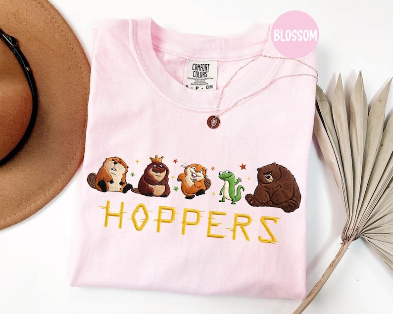 May include: A light pink Comfort Colors crew neck t-shirt featuring embroidered cartoon animals and the word "HOPPERS" in yellow. The animals include a beaver, a bear with a crown, a squirrel, a frog, and a brown bear. A gold necklace is also visible.