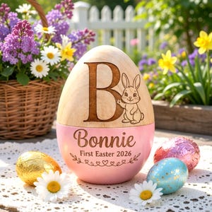 May include: A wooden Easter egg with a pink base, engraved with the name "Bonnie" and "First Easter 2026". The top features a large letter "B" and a bunny illustration. Decorative Easter eggs and flowers surround the egg.