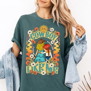 Puede incluir: Una camiseta de color verde azulado oscuro con un estampado gr&aacute;fico de dos figuras con cabello rubio y rojo que llevan sombreros de vaquero, rodeadas de flores, llamas y un sol. El texto "COWBOY LIKE ME" aparece en una fuente estilizada y llamativa.