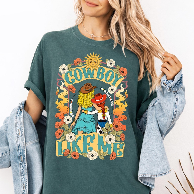 Puede incluir: Una camiseta de color verde azulado oscuro con un estampado gr&aacute;fico de dos figuras con cabello rubio y rojo que llevan sombreros de vaquero, rodeadas de flores, llamas y un sol. El texto "COWBOY LIKE ME" aparece en una fuente estilizada y llamativa.